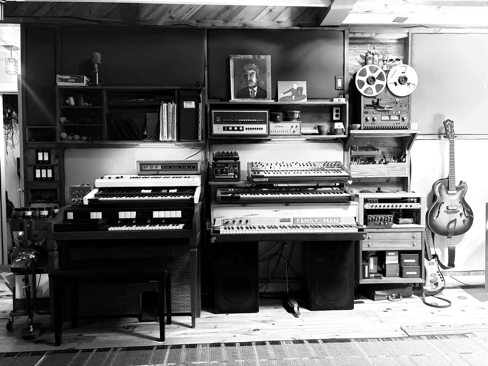 Studio wall lined with vintage keyboards, a Rhodes piano, organ, Sequential synth, reel-to-reel tape machine, and a hollow-body guitar.