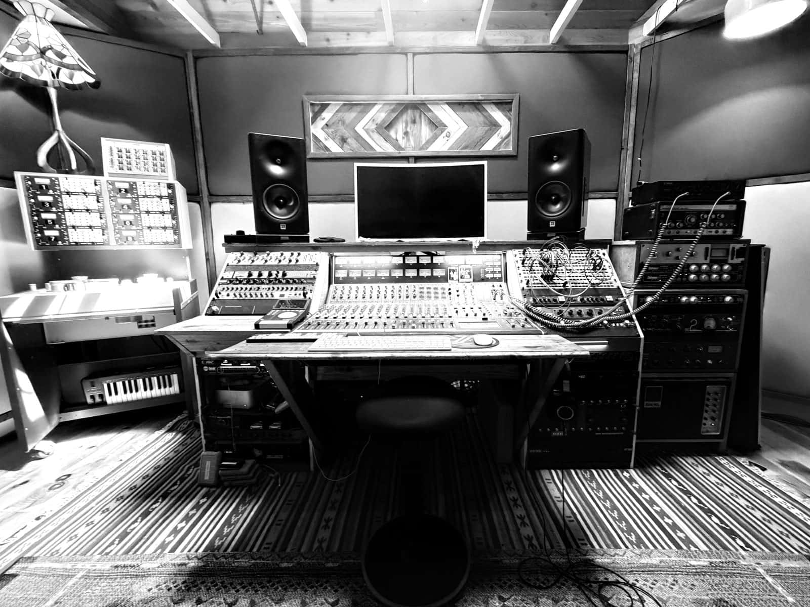 Recording studio control room: analog mixing console centered between two large monitor speakers, flanked by racks of outboard gear, with a chevron wood panel above and patterned rugs on the floor.