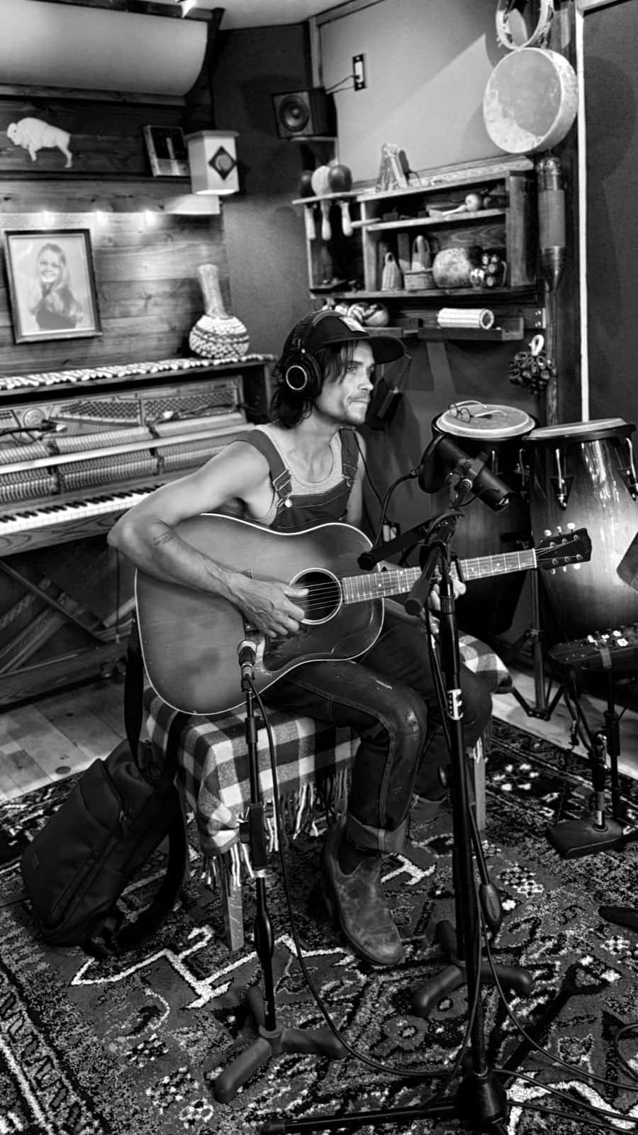 A musician in overalls and headphones plays acoustic guitar in front of a microphone, surrounded by an upright piano, congas, and shelves of percussion.